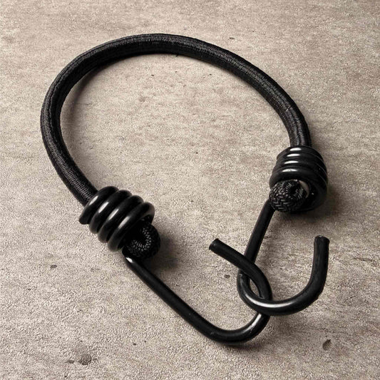 A black metal bungee hook with a textured wrap on both ends, resting on a textured gray concrete surface.