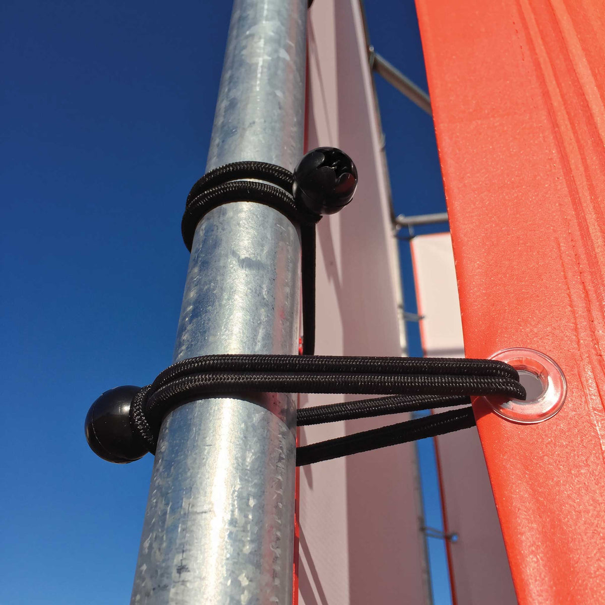 Black bungee cords with spherical black end caps securing a red fabric panel to a galvanized metal pole, photographed outdoors against a clear blue sky.
