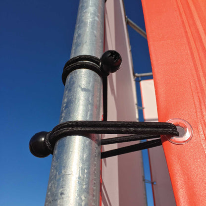 Black bungee cords with spherical black end caps securing a red fabric panel to a galvanized metal pole, photographed outdoors against a clear blue sky.
