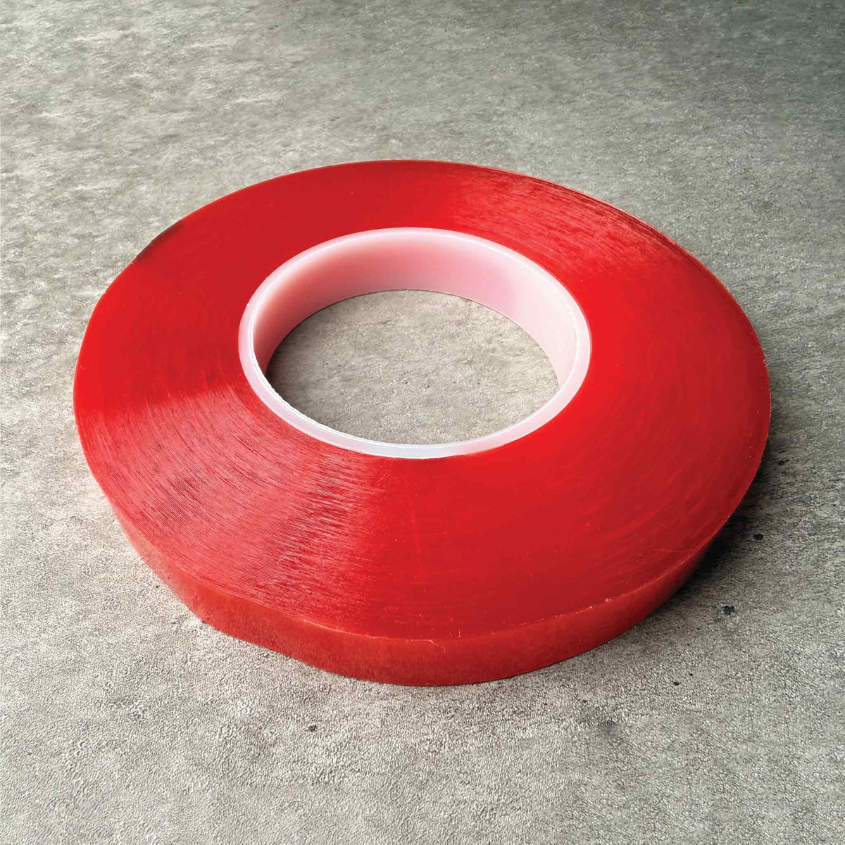 A roll of double sided adhesive tape with a , photographed on a textured gray concrete surface.
